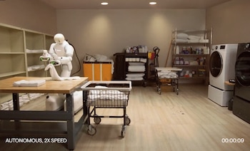 Un robot humanoide blanco sostiene una toalla de color blanco sobre una mesa de madera, rodeado por cestos de ropa y electrodomésticos