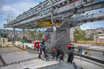 Obreros con cascos trabajan junto a una cabina cubierta bajo la estructura metálica de una estación de teleférico