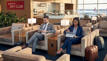 Dos personas en una sala VIP de aeropuerto: un hombre con celular y una mujer con tableta, sentados en sofás junto a equipaje con aviones al fondo