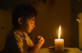 Un niño con camisa clara, ojos cerrados y manos unidas, sentado junto a una vela blanca encendida que irradia luz amarilla en un cuarto oscuro