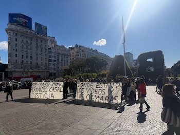 Personas marchan en una plaza soleada sosteniendo pancartas blancas con texto negro que pide justicia para Fernando Casal. Edificios clásicos de fondo