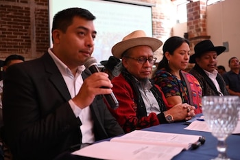 Un hombre con traje y camisa blanca sostiene un micrófono, junto a tres personas sentadas en una mesa con documentos, un vaso y una pantalla