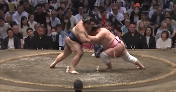 Dos hombres luchadores de sumo en un cuadrilátero de tierra, rodeados por una multitud de espectadores sentados, con un árbitro de pie al fondo