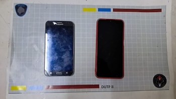 Dos teléfonos móviles, uno negro Samsung a la izquierda y otro con funda roja a la derecha, sobre una superficie gris con patrón de cuadrícula y logos policiales