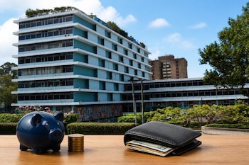 Una alcancía azul, una pila de monedas y una billetera abierta con billetes sobre una mesa de madera, frente a un edificio de varios niveles.