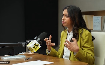 Mujer con blazer verde lima gesticula en un estudio. Un micrófono de "The Mel Robbins Podcast", un libro y un vaso de agua son visibles sobre la mesa de madera