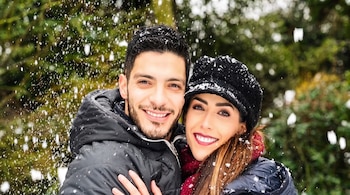 Hombre y mujer sonrientes con ropa de invierno, rodeados de nieve que cae y árboles verdes borrosos al fondo.
