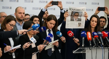 Una docena de periodistas con micrófonos y cuadernos en una sala. Varios sostienen teléfonos para grabar, y un hombre al fondo levanta un periódico visible.