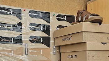 Una sandalia marrón y blanca sobre cajas de cartón. Al fondo, un tablero de corcho con patrones de calzado de la marca Viamo y etiquetas de colores