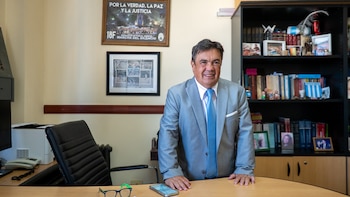 Hombre con traje gris y corbata azul sonríe detrás de un escritorio de madera. Detrás, una estantería con libros y objetos, y a la izquierda una silla de oficina