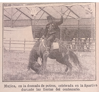 Un jinete con sombrero oscuro y ropa de montar domando un caballo en un corral de tierra, con una cerca baja y una estructura techada al fondo