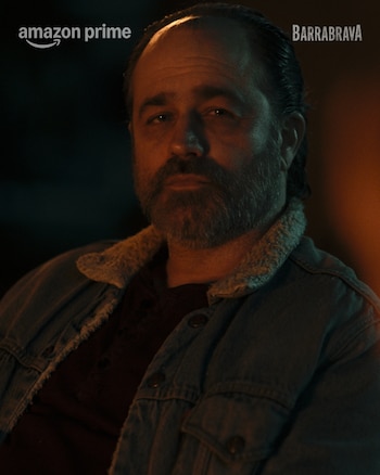 Hombre de mediana edad con barba y cabello oscuro, vestido con una chaqueta de mezclilla con cuello de borrego, mirando al frente en un entorno tenue y oscuro