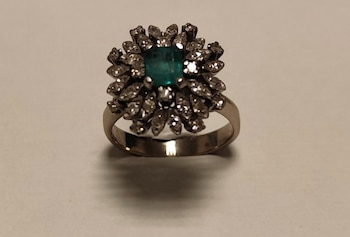 Un anillo metálico con una piedra verde cuadrada en el centro y pequeños brillantes alrededor sobre un fondo claro
