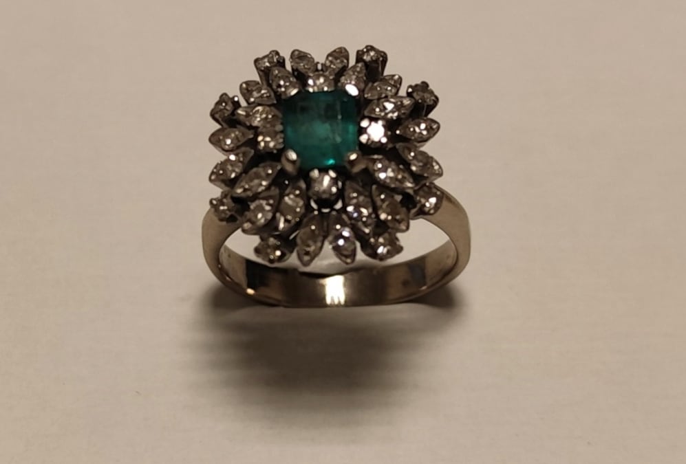 Un anillo de compromiso presenta una piedra verde de corte rectangular rodeada por múltiples gemas brillantes en forma de flor