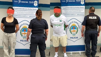 Una mujer y un hombre con los ojos cubiertos permanecen de pie junto a policías frente a un cartel institucional
