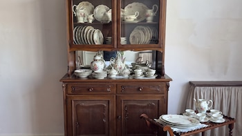 Mueble de madera con vitrina, repisas y cajones, lleno de vajilla blanca con flores, junto a un carrito de servicio con tazas y platos.