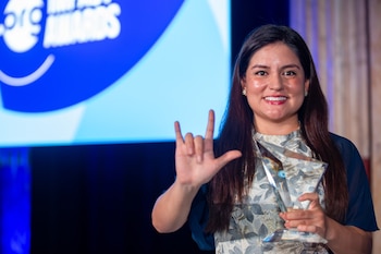 Una mujer sonriente sostiene un trofeo de vidrio y levanta la mano con los dedos extendidos frente a una pantalla azul