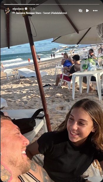 Primer plano de un hombre tatuado sonriendo junto a una niña. Detrás, una sombrilla, arena, el mar con olas y personas