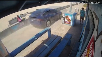 Un automóvil azul circula sobre un paseo peatonal junto a la playa y levanta polvo cerca de un puesto de comida y un transeúnte