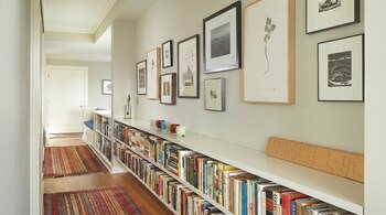 Pasillo interior con paredes claras, estanterías blancas llenas de libros y arte enmarcado. Suelo de madera con alfombra decorativa rojiza.