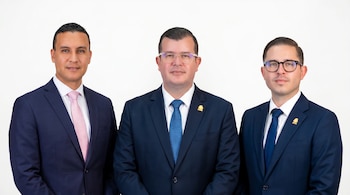 Una foto de medio cuerpo de tres hombres con trajes azul marino, camisas blancas y corbatas, posando formalmente frente a un fondo blanco liso.