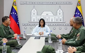 Una mujer con gafas y blusa clara sentada a una mesa de reunión, rodeada de cinco hombres con uniformes militares. Dos banderas de Venezuela a los lados