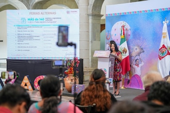 Una mujer con vestido estampado y cabello oscuro habla desde un podio, flanqueada por las banderas de México y Oaxaca, frente a una pantalla con la agenda de la Guelaguetza 2026