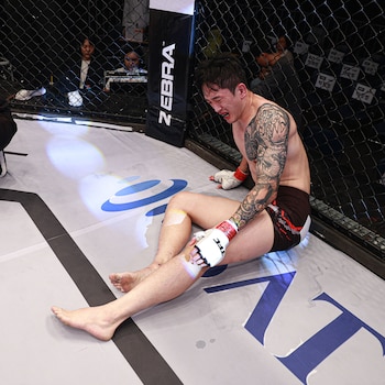 Hombre con guantes y tatuajes en el brazo izquierdo sentado en la lona de una jaula deportiva con la boca abierta y gesto de dolor