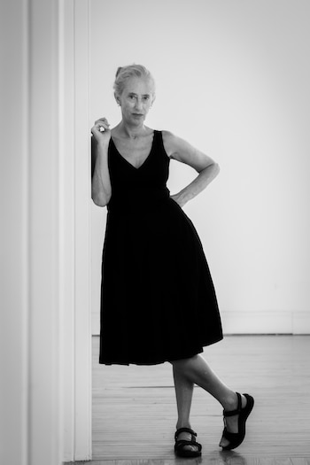 Una mujer de cabello cano recogido, con un vestido negro y sandalias oscuras, se apoya en una pared blanca lisa frente a un piso de madera