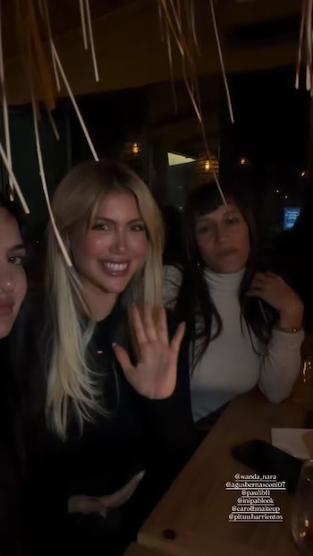 Wanda Nara sonríe y saluda con la mano, sentada entre dos mujeres en un ambiente de restaurante con luz tenue y adornos colgantes