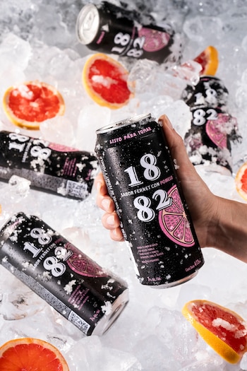Una mano sostiene una lata negra de 1882 sabor fernet con pomelo, cubierta de hielo y gotas, entre otras latas similares, hielo y pomelos partidos