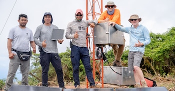 Cinco hombres de pie junto a una estructura metálica naranja con paneles solares y cajas técnicas, rodeados de vegetación