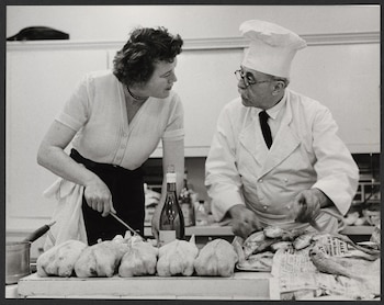 Mujer de cabello oscuro con un chef de gorro blanco y gafas en una cocina. Frente a ellos, aves de corral, pescado y una botella de vino