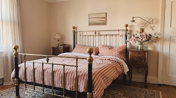 Um quarto com cama cobre e preta, colchas brancas e vermelhas, duas mesas de cabeceira, candeeiros, quadros, tapetes e cortinas nas janelas.