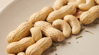Cacahuates con cáscara y nueces de la India peladas mezclados sobre una superficie beige clara con algunos restos de hierbas verdes.