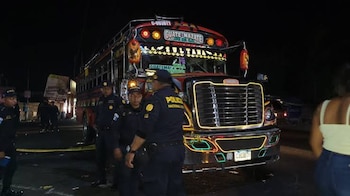 Autobús rojo con luces LED y texto "GUATE-MAZATE", cinco agentes de policía con uniforme azul, una mujer de espaldas, cinta amarilla en el suelo, de noche