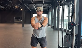 Un hombre usa una máquina de poleas en un gimnasio. Lleva gorra, camiseta sin mangas y pantalón corto. Se ven paredes oscuras y ventanas