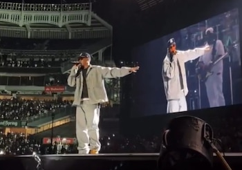 Un hombre con gorra, gafas y ropa clara canta en un escenario. Detrás, una pantalla gigante muestra al artista y público en las gradas de un estadio nocturno