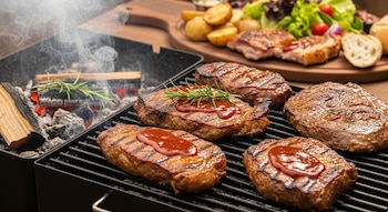 Una parrilla con trozos de carne asada, salsa barbacoa, romero, brasas encendidas, astillas de madera y humo. En segundo plano, guarniciones.