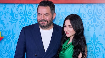 Un hombre con barba, saco azul y camiseta blanca posa junto a una mujer con vestido verde frente a una pared azul decorada.