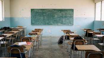 Interior de aula con pizarrón verde, paredes blancas y azules desgastadas. Filas de pupitres de madera y metal, algunos con mochilas y cuadernos dispersos.