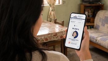 Una mujer vista desde atrás, con cabello oscuro, sostiene un smartphone que muestra la página de resultados de la ONPE con los votos de Keiko Fujimori. Se ve una mesa y una habitación