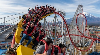 Grupo de personas en una montaña rusa amarilla y roja con asientos giratorios, Eejanaika, con expresiones de emoción y el Monte Fuji nevado al fondo.