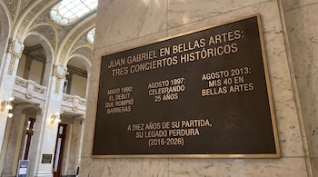 Placa metálica en una columna de mármol con texto conmemorativo sobre Juan Gabriel, con arcos y pasillos del recinto al fondo.