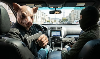 Dos personas dentro de un coche. El conductor lleva una máscara de cerdo y una pistola. El pasajero usa pasamontañas y sostiene otra pistola