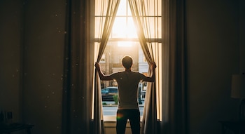 Persona de espaldas frente a una ventana con cortinas abiertas. Luz solar brillante entra por la ventana. El interior de la habitación está oscuro.