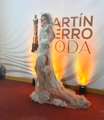 Mujer rubia en vestido largo blanco translúcido con volantes sobre alfombra roja, posando frente a un cartel de los Martín Fierro y una estatuilla dorada