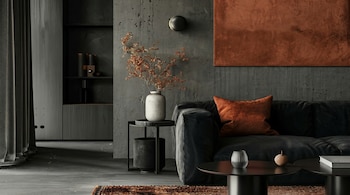 Vista de una sala de estar moderna con paredes de concreto gris oscuro, un sofá de terciopelo negro, un cojín naranja y una obra de arte cobriza en la pared.