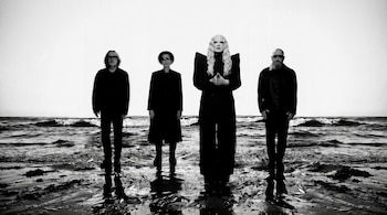 Fotografía en blanco y negro de cuatro personas vestidas de negro de pie sobre la orilla mojada frente al mar