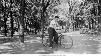 Fotografía en blanco y negro de un joven sentado sobre una bicicleta en un camino de tierra rodeado de árboles.